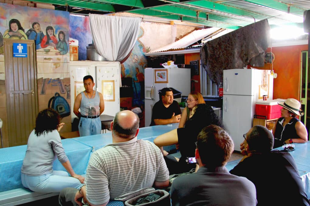 A group of people, some appearing White and some Hispanic, are in a makeshift meeting space, sitting at tables, apparently in discussion. There are bright murals on the wall. The figure speaking in front is a middle-aged Hispanic man, wearing a silver cross around his neck. 