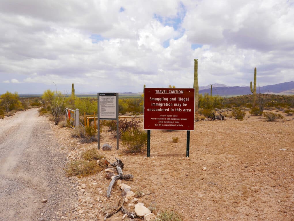Color photo of gravel road in desert, saguaro cacti visible, and large sign that says "Travel Caution. Smuggling and Illegal Immigration may be encountered in this area."