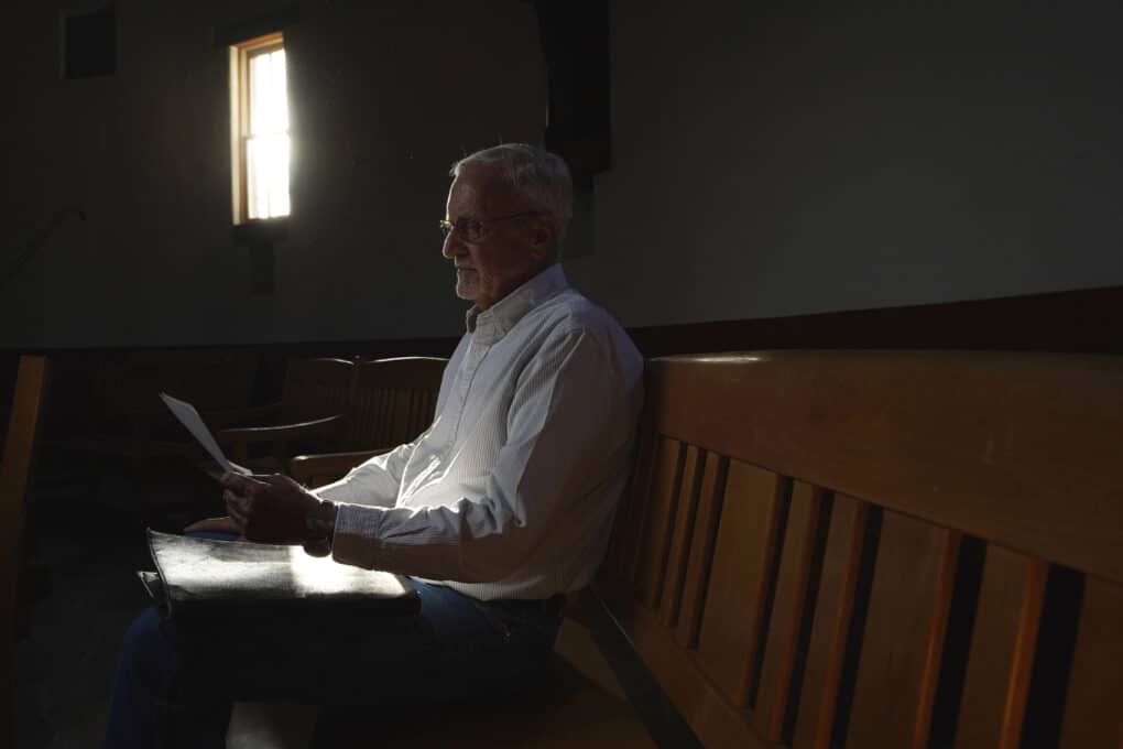 An older white man in a collared white shirt is looking at papers in his lap; sun streams in through a window. 