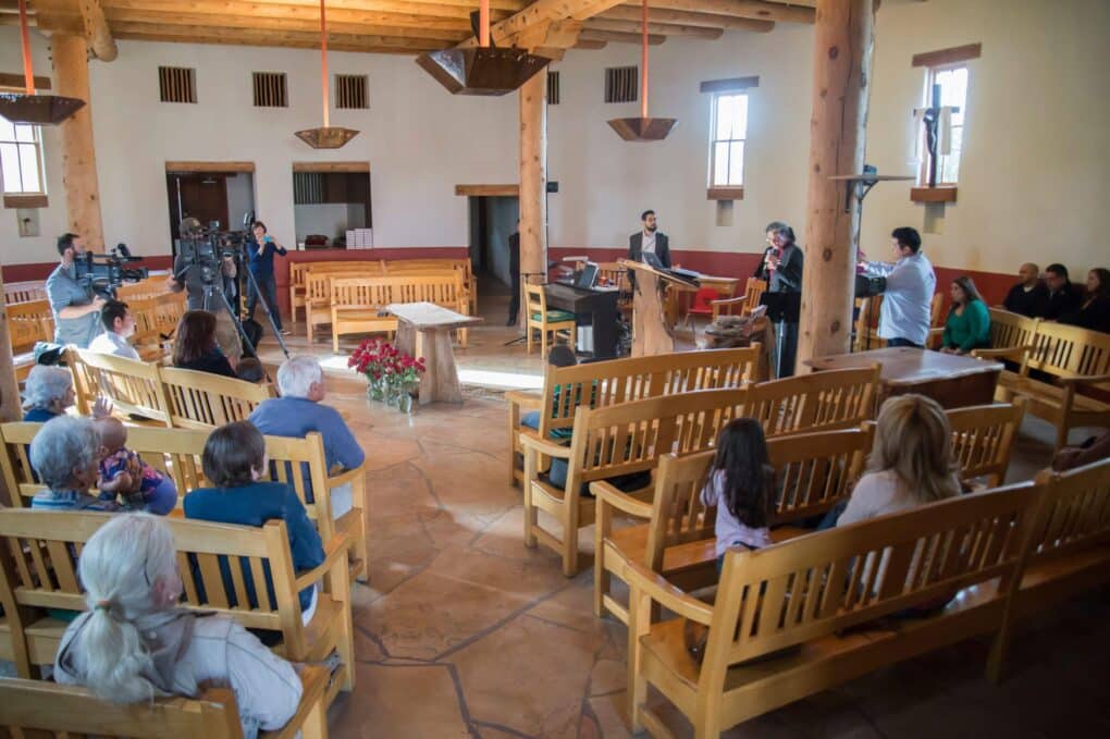 Interior of church with light wood benches arranged in a circular fashion around pulpit where a woman is speaking to a small audience with television cameras. 