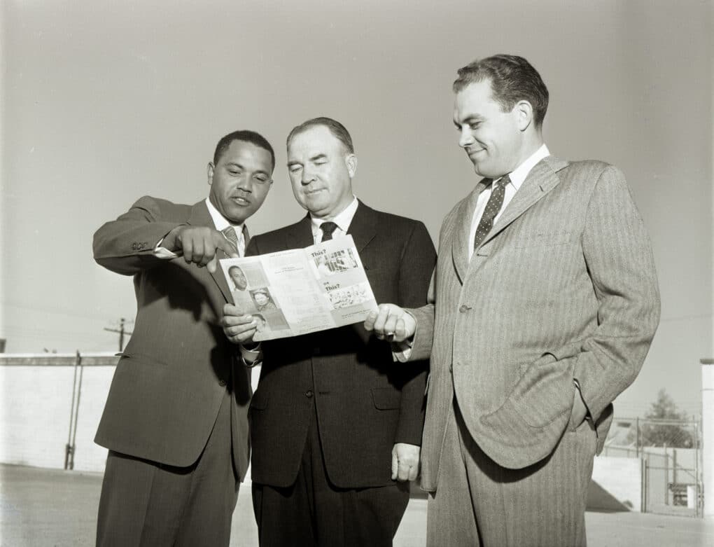 Black and white photo of three men in suits, smiling and looking at a brochure together. Photo seems staged, man on left is Black and the others are older and White.