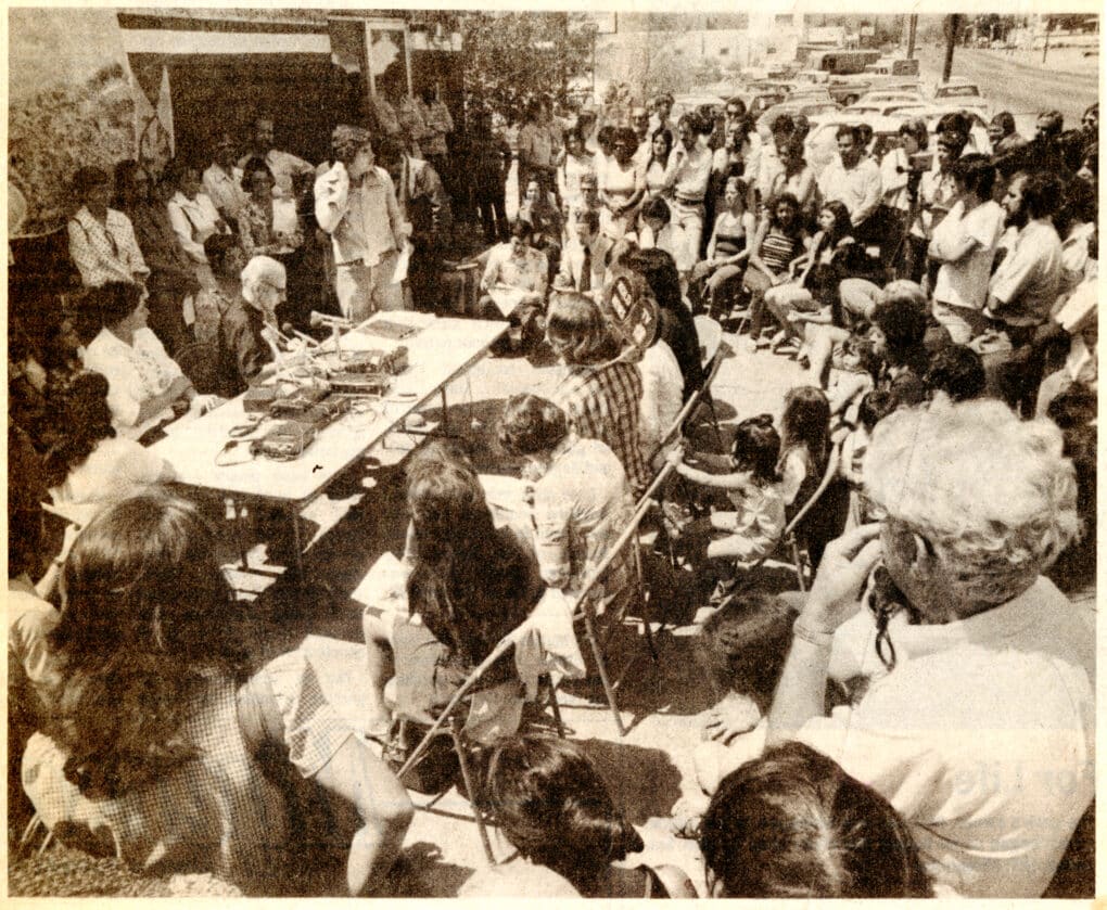 Black and white photo showing crowded scene with many people standing and sitting outdoors in folding chairs, listening to a speaker sitting at table with microphone.
