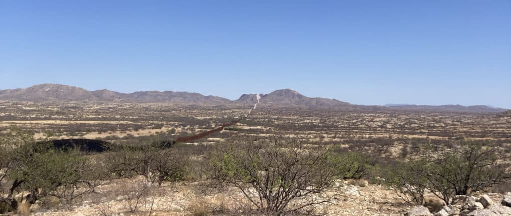 Landscape photograph of Arizona desert; a small mountain rises in the far distance and the iron border wall is visible.