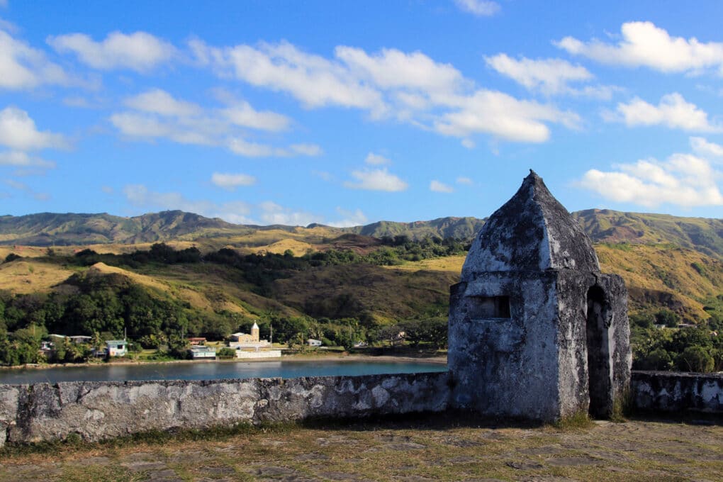 Color photograph of stone parapet and guardhouse overlooking a bay, with a church and green hills on the opposite shore.