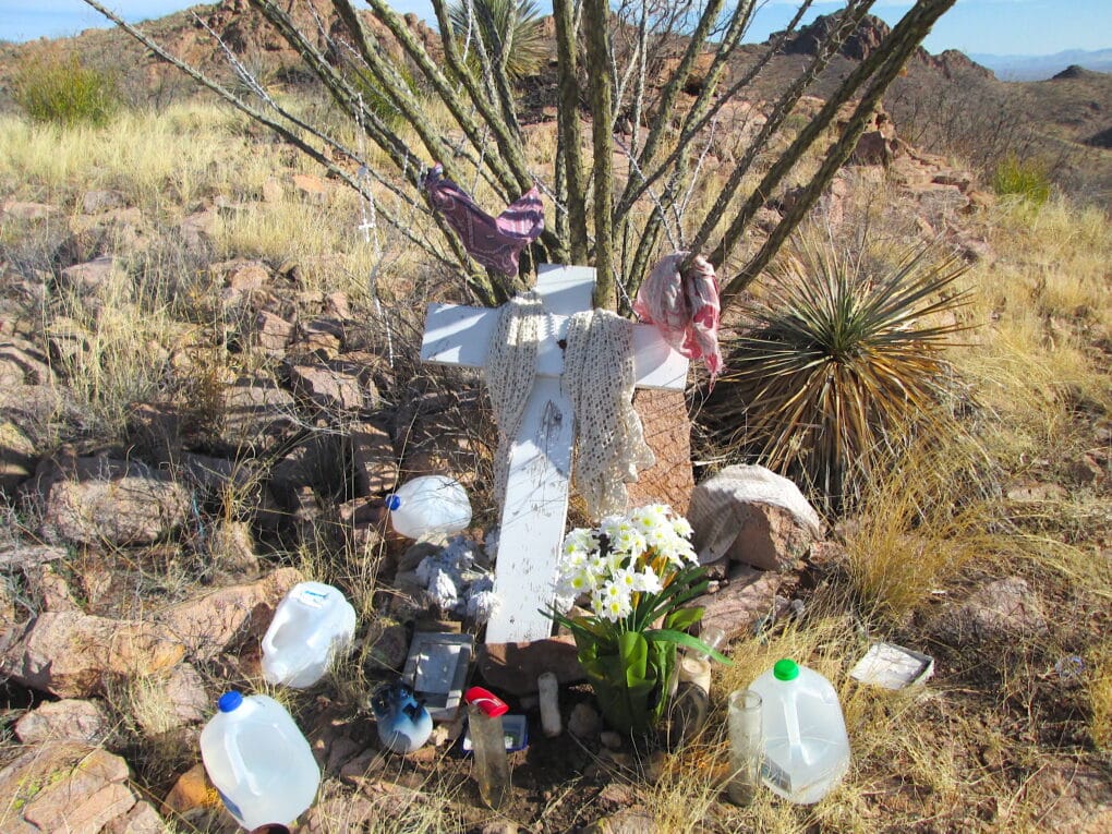 A white cross is perched against a desert bush, surrounded by plastic jugs filled with water and some scarves and flowers.
