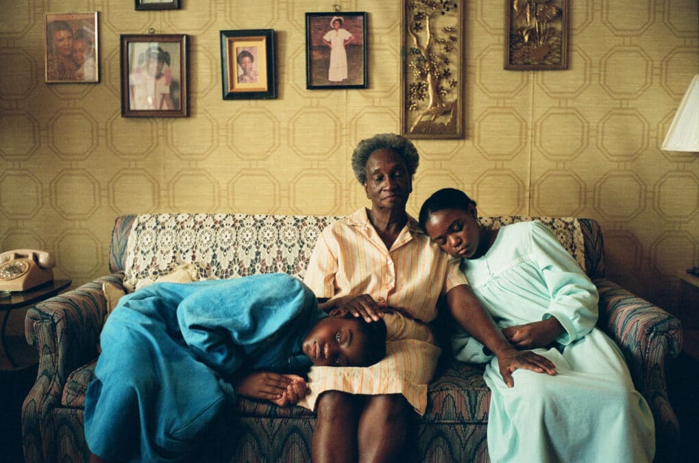 Color film still of elderly Black woman on a sofa in a domestic interior, with two Black girls seated beside her.
