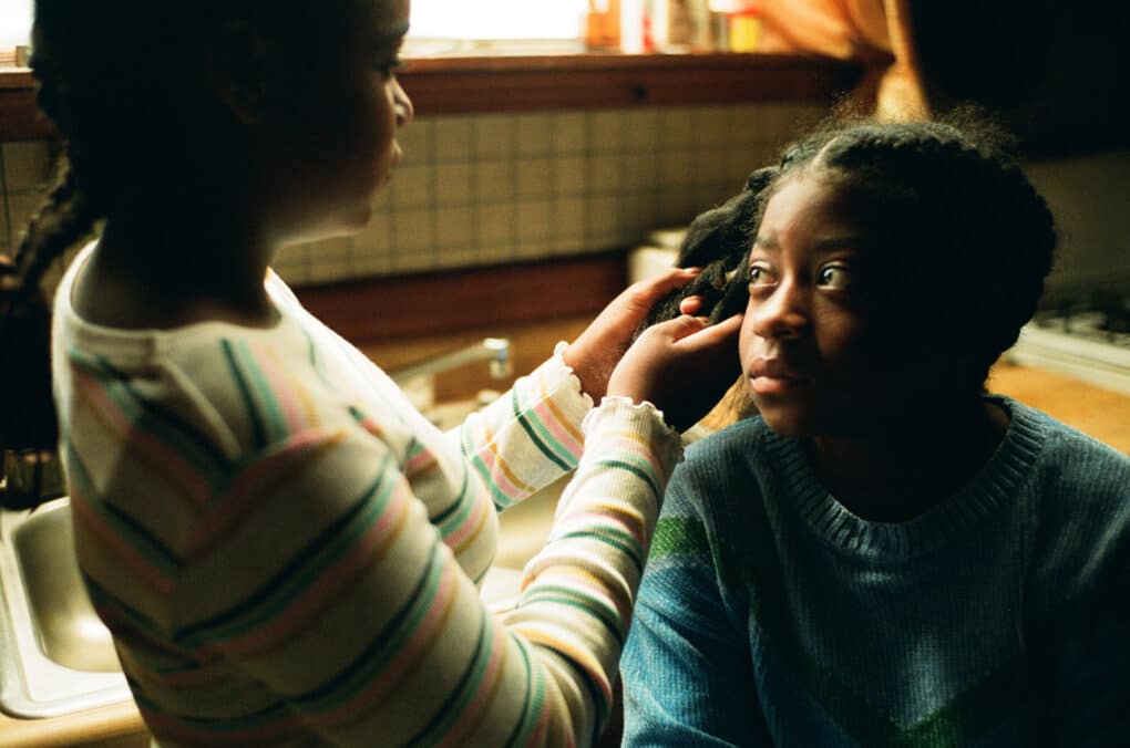 Color film still of one Black girl braiding another's hair in a 1970s-style kitchen.
