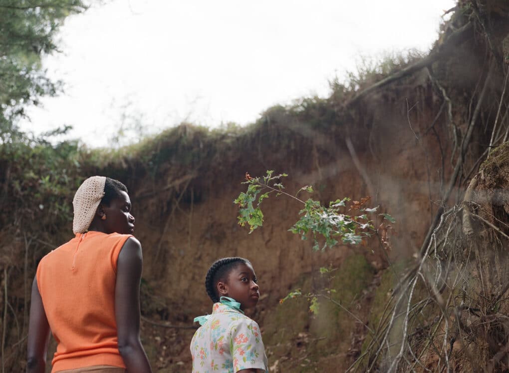 Color film still of Black woman and Black girl standing in front of a tall bank of clay.