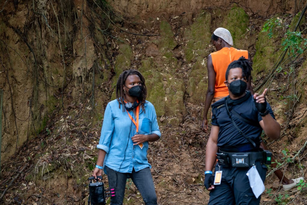 Color production still of two Black women and a Black man--film director, actor, and cinematographer--standing in front of a high clay bank.