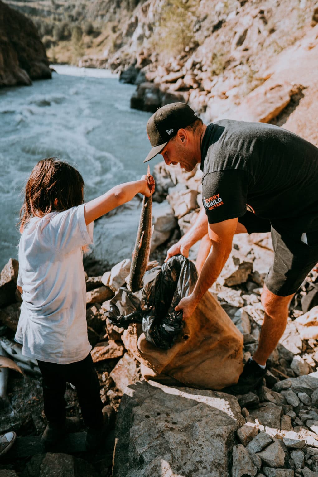 Color photograph of young man in baseball cap, shorts, and t-shirt with child in t-shirt, putting fish into a bag beside fast-moving river.