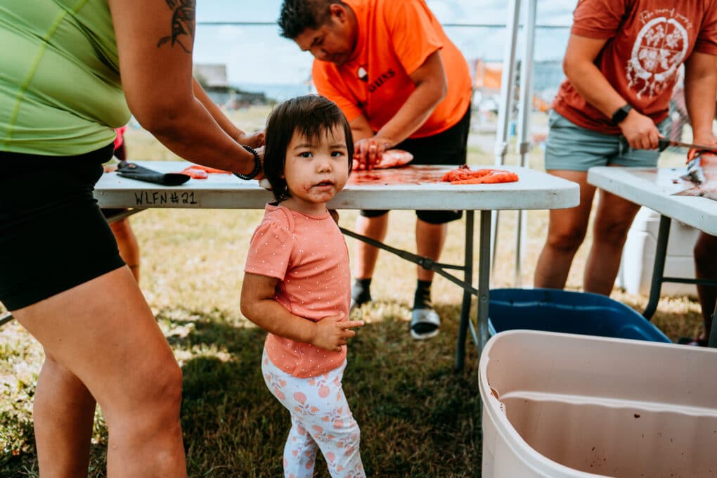 Color photograph of three adults in t-shirts and shorts filletting salmon on folding tables outdoors; in the foreground, a small child with fishblood on her mouth looks directly at the camera.