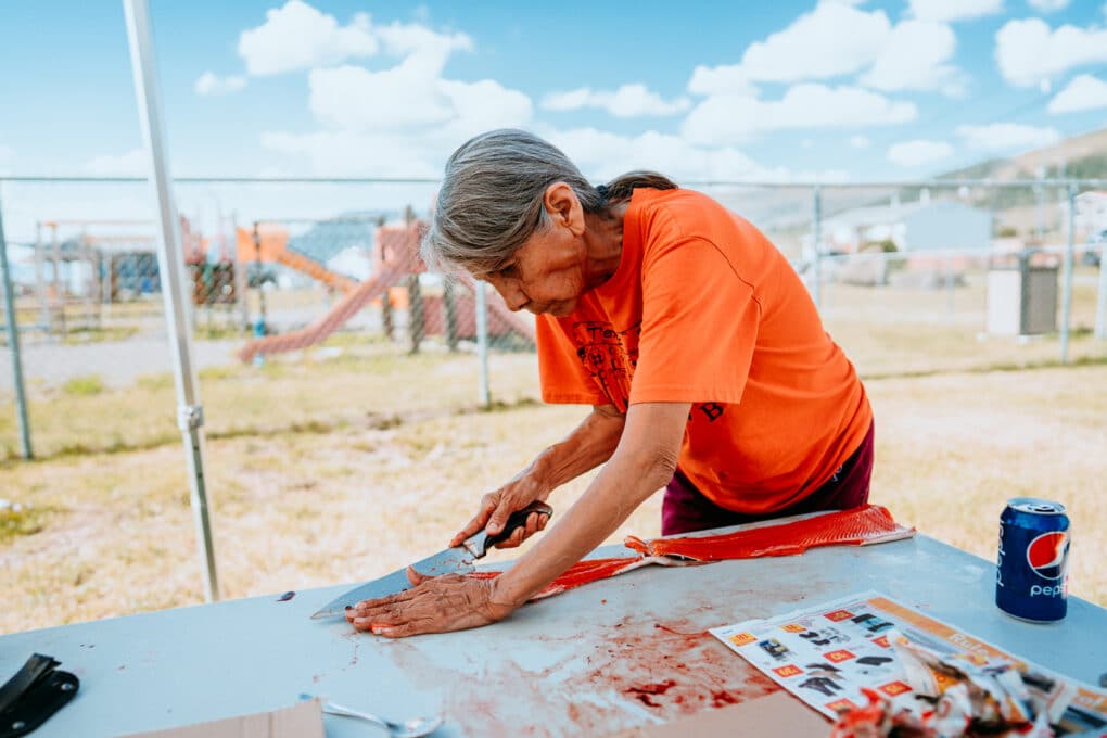 Color photograph of gray-haired woman in orange t-shirt filleting salmon at an outdoor folding table. In the background is playground equipment behind a chainlink fence.