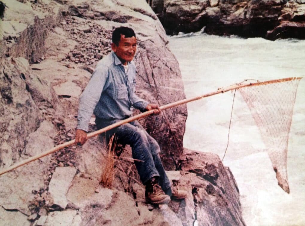 Color photograph of elderly dark-haired man propped on rocks above fast-flowing waters, holding a large net on a long pole and smiling at the camera.
