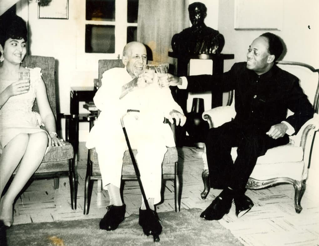 Three people are sitting in chairs, smiling, and toasting drinks. On the left is a woman with a bouffant hairdo, in the center an older man with a cane resting on his leg, to the right, clinking glasses with the other man, is a man dressed in a black suit.