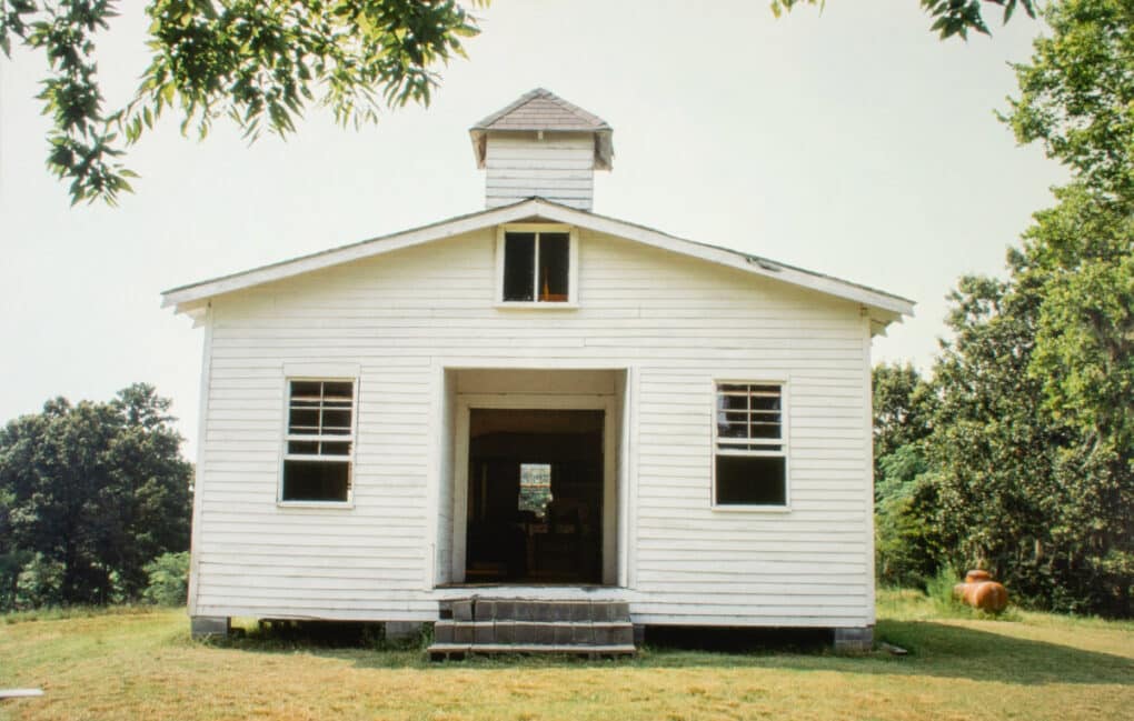 Color photograph of white clapboard church in grassy space surrounded by green trees.