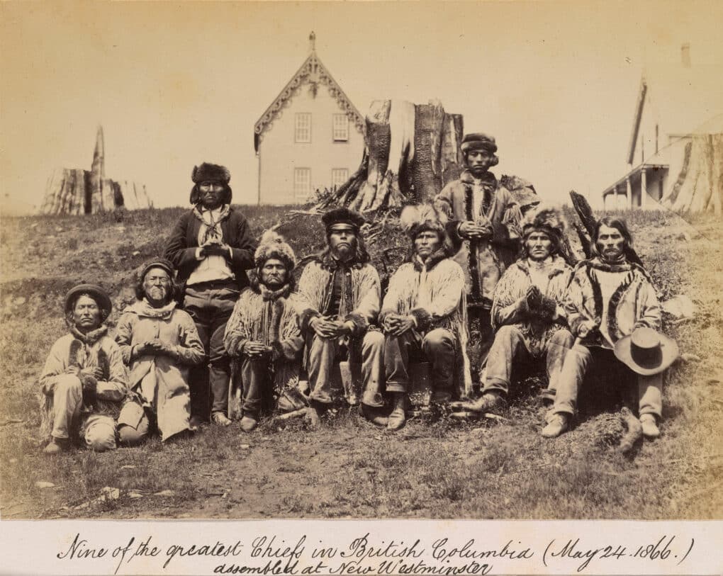 Sepia-toned photograph of nine Indigenous men posing for the camera with two wooden houses in the background.