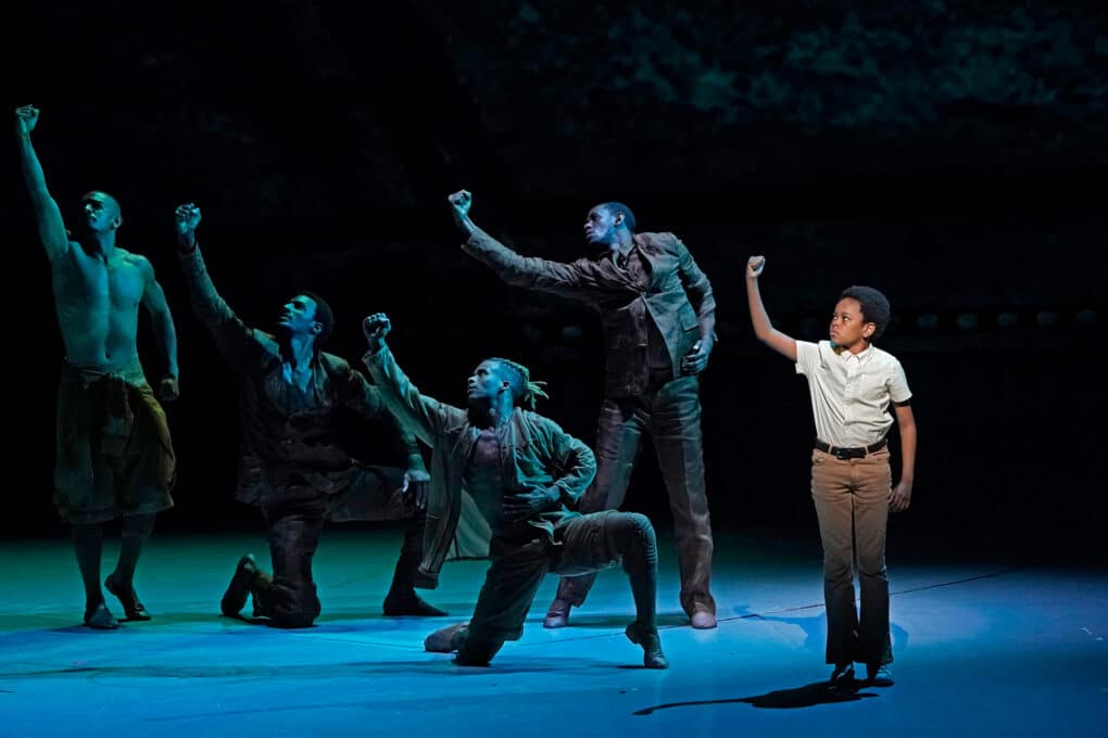 Color photograph from stage production: Four young Black male dancers in shadow and one Black boy in spotlight, all raising their right fists.