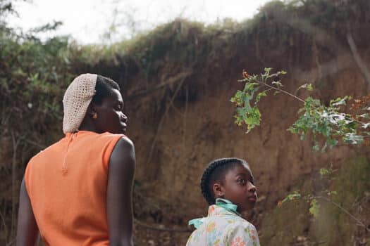 Color film still of Black woman and Black girl standing in front of a tall bank of clay.