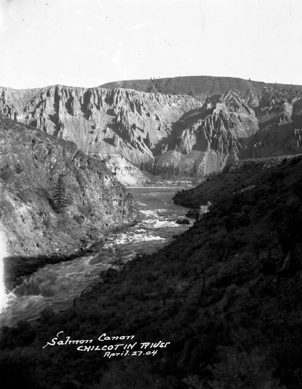 Black-and-white photograph of river flowing through badland cliffs; caption handwritten in white at lower edge.