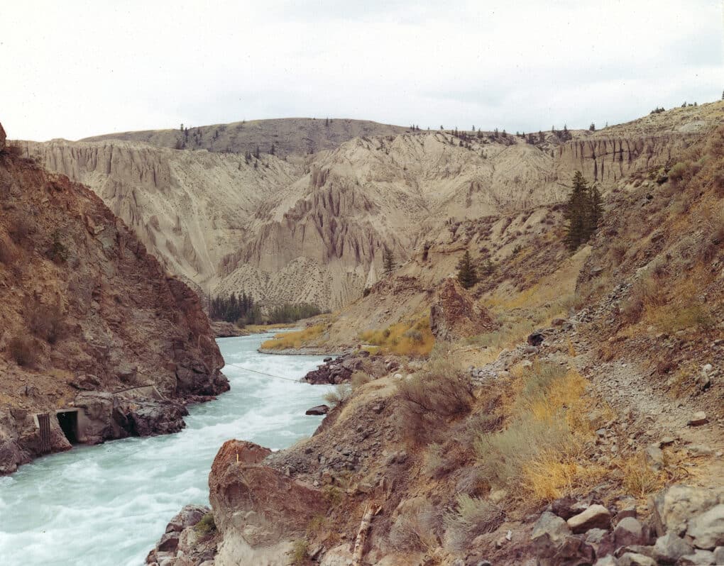 Color photograph of fast-flowing blue-tinged river flowing through red badland cliffs.