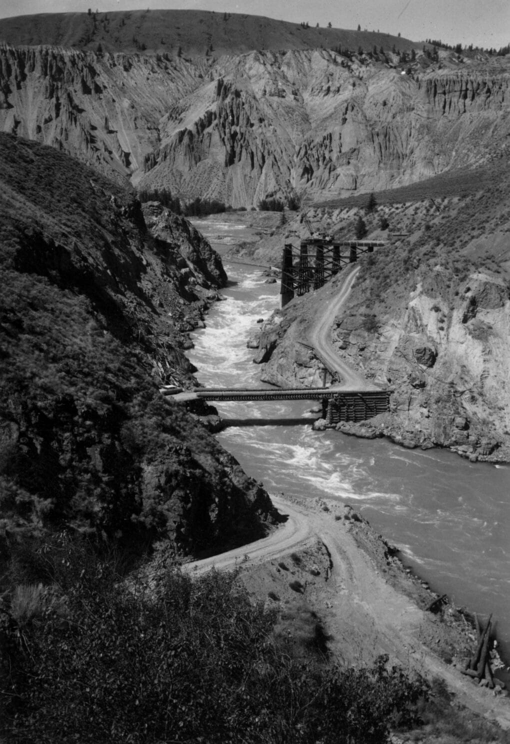 Black-and-white photograph of river bridge under construction upriver from another, smaller bridge.