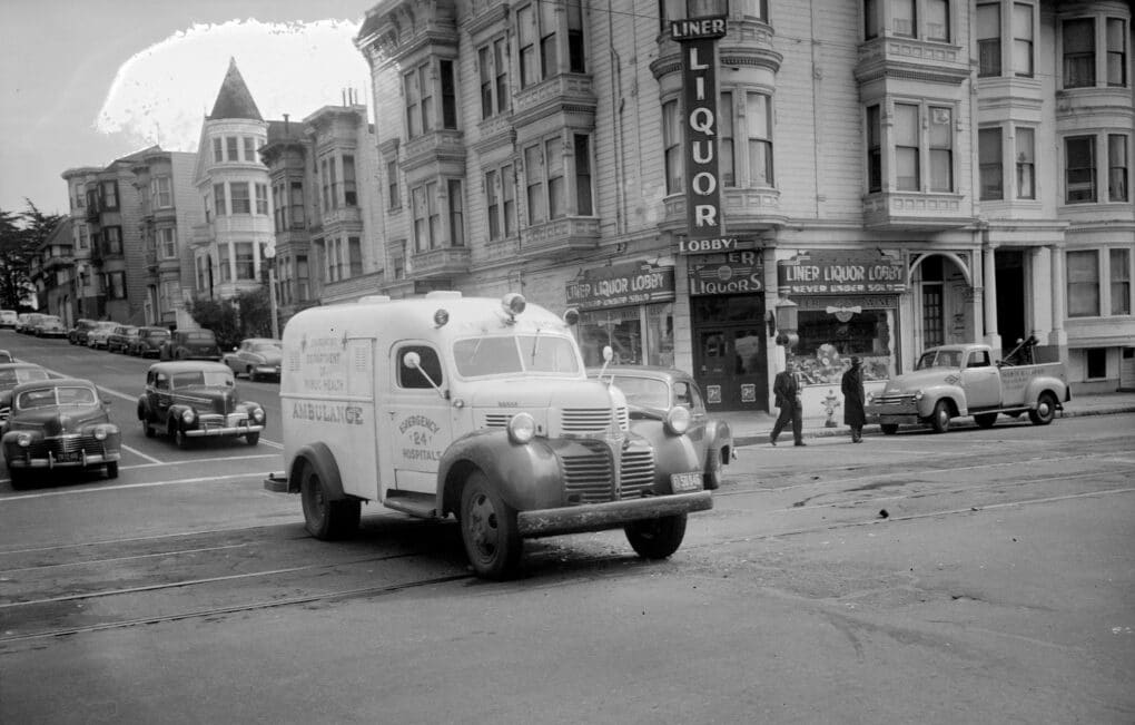 Black-and-white photograph of midcentury ambulance crossing an intersection beside tall wooden Victorian buildings, one bearing a sign reading LIQUOR.