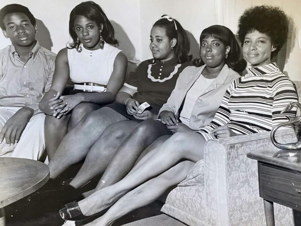 Black-and-white photograph of Black family of five (one teenage boy, three teenage girls, and young mother) all in early 1970s clothes, seated on a sofa in a domestic interior.