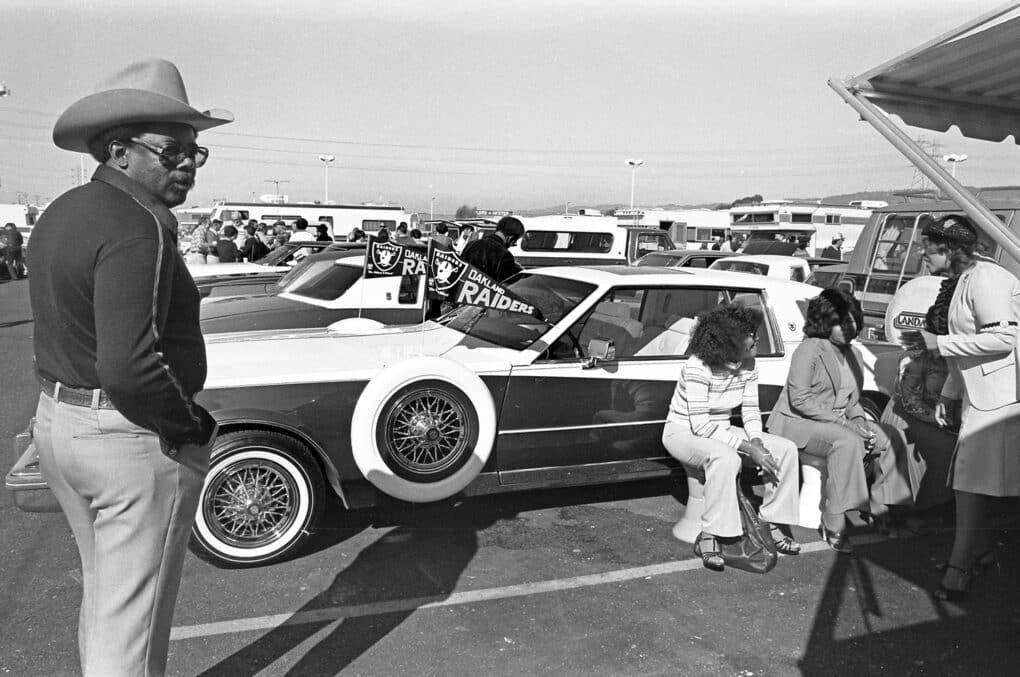 Black and white photograph of tall Black man in cowboy hat, along with several women in a parking lot; a large car is flying OAKLAND RAIDERS flags.