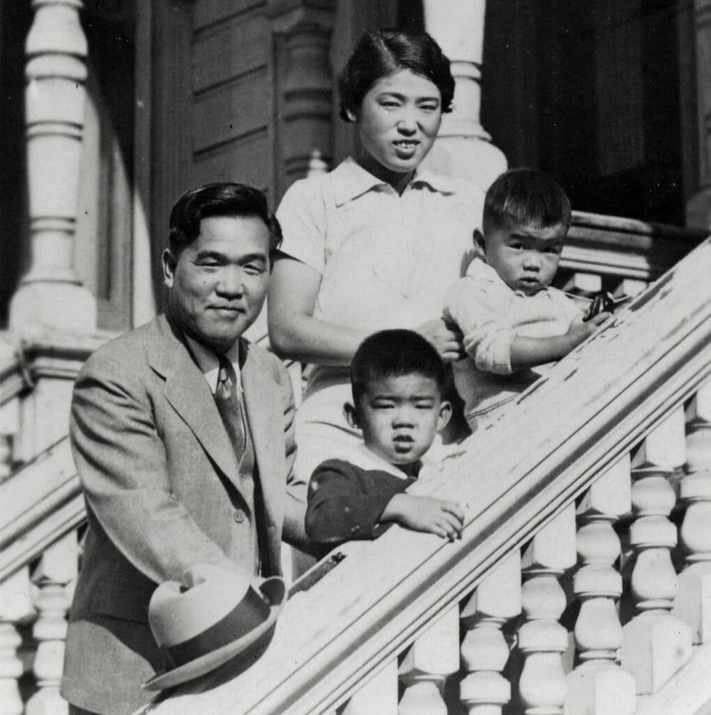 Black-and-white photograph of Japanese family (father, mother, and two small children )on front steps of Victorian house.