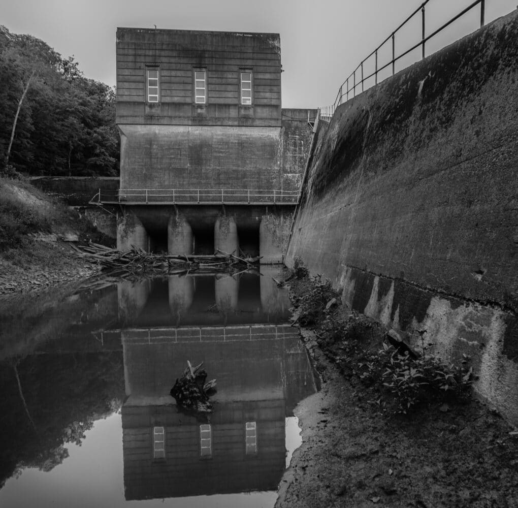 Black and white photograph of a river running through sluice gates. Branches and other debris hinder the flow of water into the gates. On the right side of the river is a shallow muddy bank.