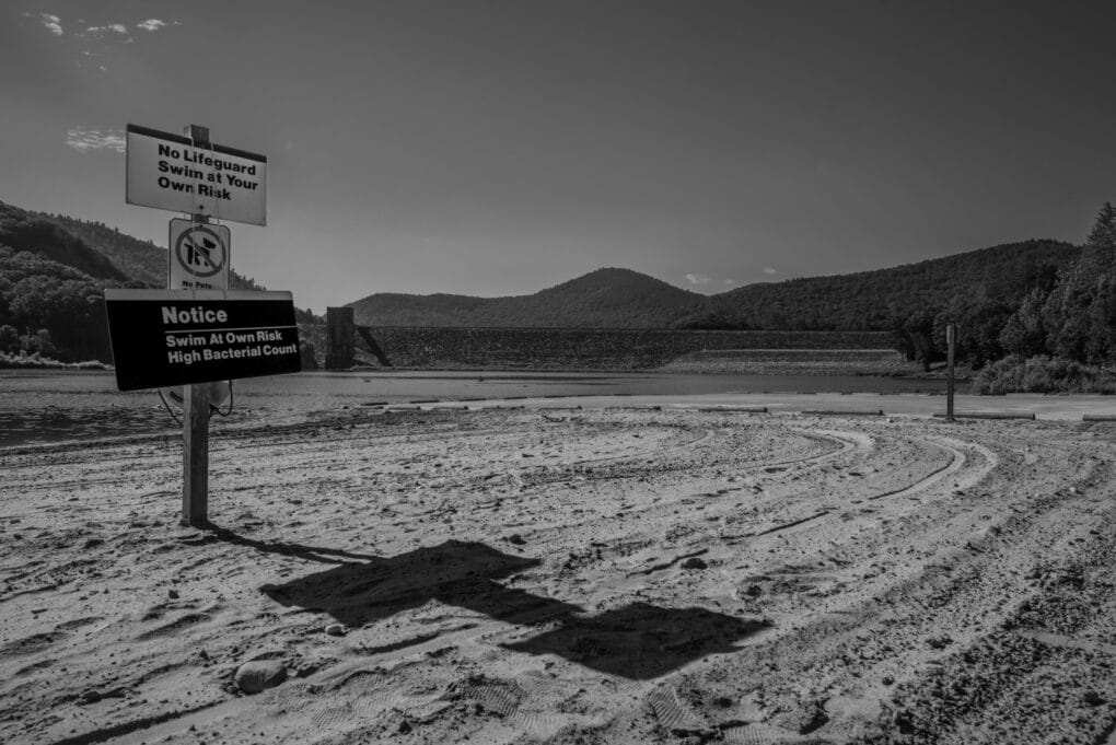Black and white photograph of a sandy plain. In the distance is a dam wall. Two signs in the foreground advise visitors to "swim at your own risk."