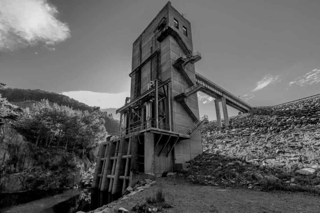 Black and white photograph looking up from the base of an intake tower. A maintenance stair climbs the side of the tower.