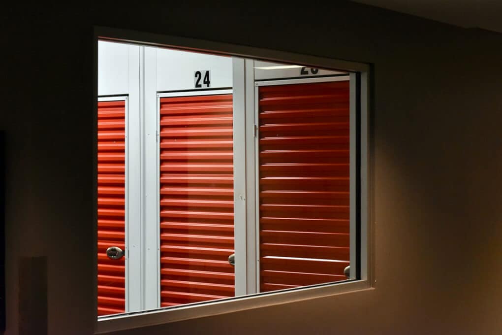 Color photograph of three red roll-up doors to storage units, seen through a glass panel from a darkened hallway. 