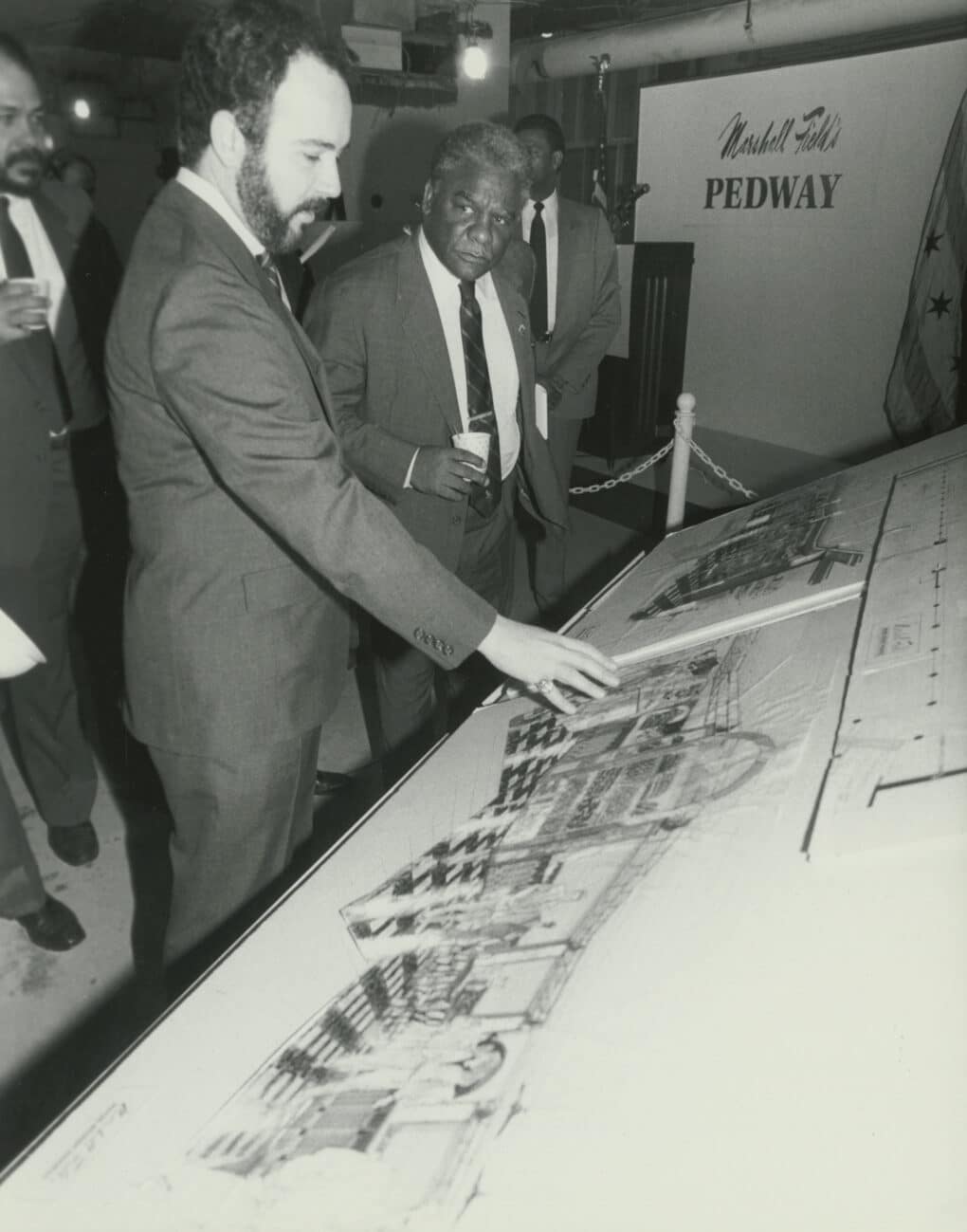 Black-and-white photograph of a Black man and a White man, both in suits, examining an architect's rendering mounted on a display shelf. In the background, wall signage reads "Marshall Field's Pedway."