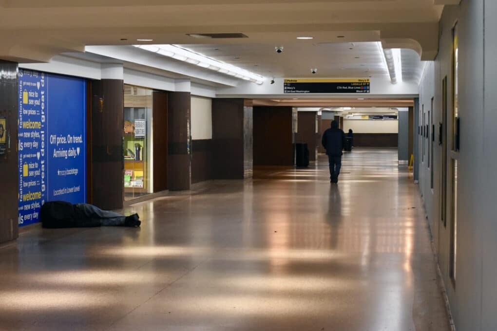 Color photograph of mostly empty underground passage way. We see one occupied retail space, one pedestrian, and a person wrapped in a blanket stretched asleep on the floor.