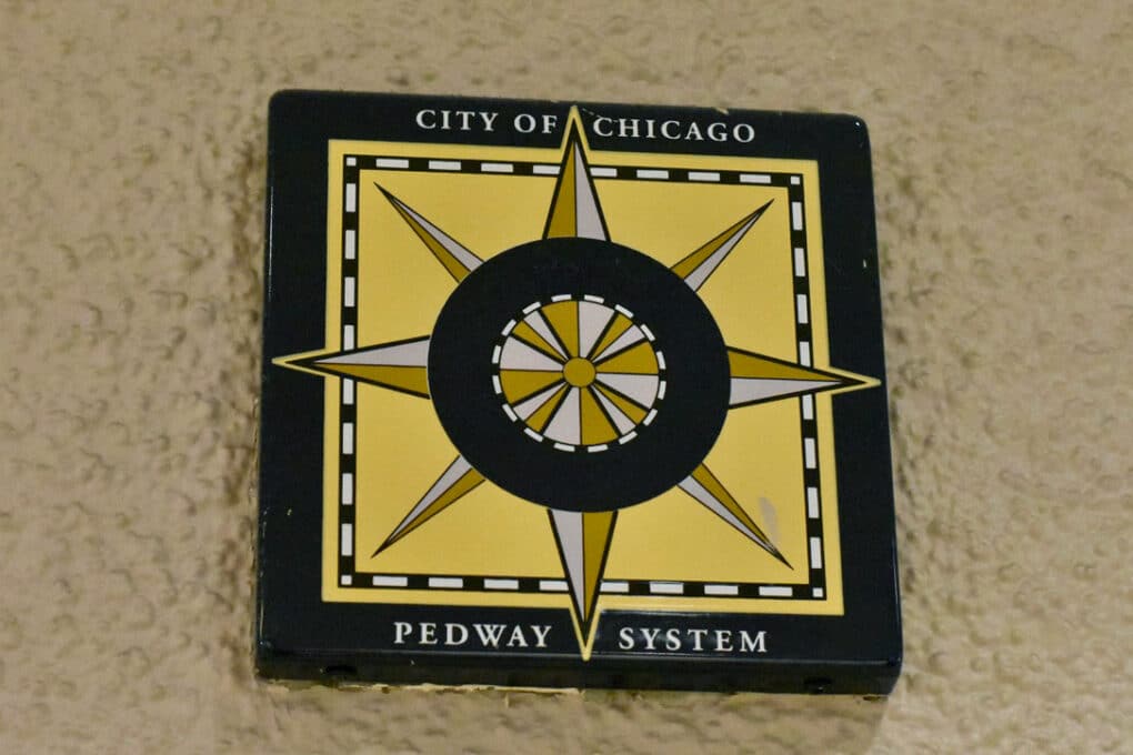 Color photograph of a yellow-and-brown compass rose labeled "City of Chicago Pedway System," mounted on a beiges stucco wall.