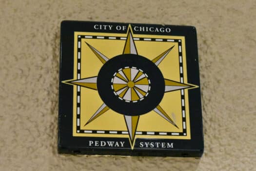 Color photograph of a yellow-and-brown compass rose labeled "City of Chicago Pedway System," mounted on a beiges stucco wall.