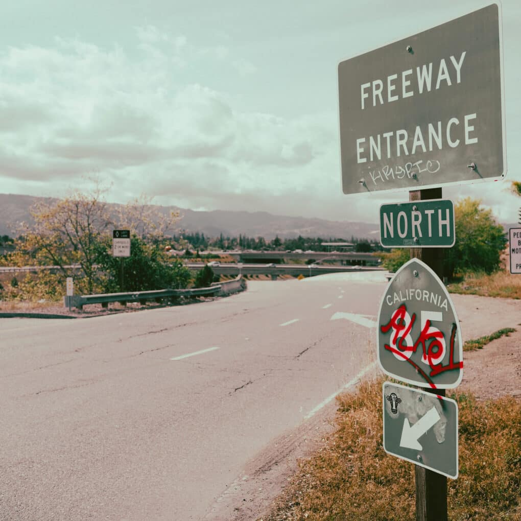 Color pinkish tone photo of freeway onramp with illegible bright red graffiti on 85 sign.