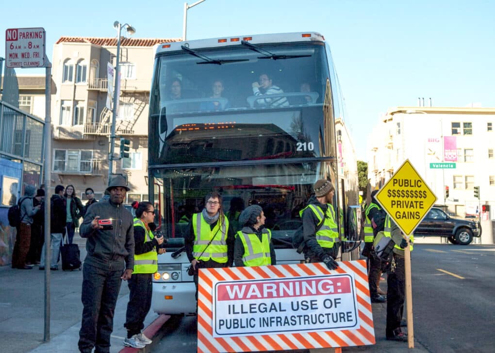 About seven people in reflective vests are blocking the path of a double decker bus that appears to be full of passengers. They have a sign that says "Warning: Illegal Use of Public Infrastructure."