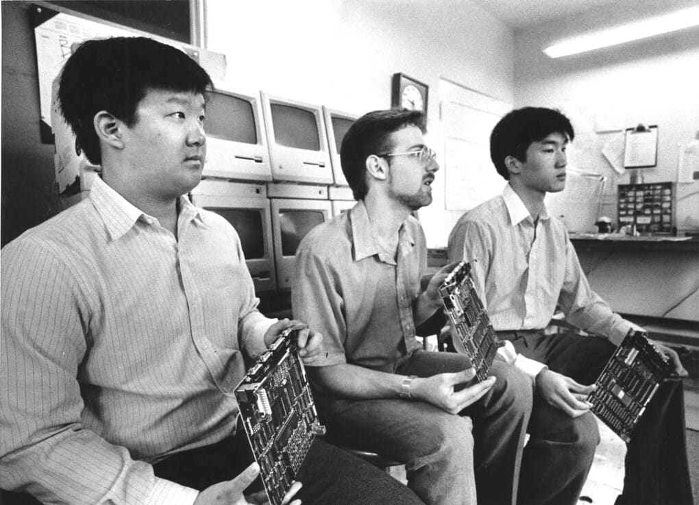 Black and white photo of three men posing with electronic motherboards. Two are Asian and one is white, behind them is a stack of Macintosh desktop computers.