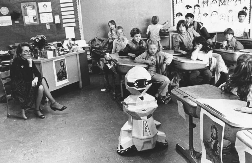 Photo of robot moving between desks in a classroom. Children are watching and laughing.