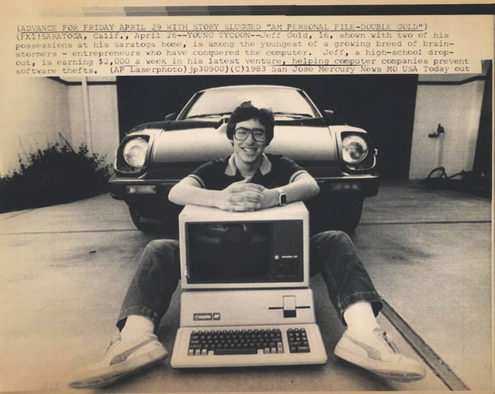 Old newspaper photo of a young man sitting in his driveway. Between his legs is a computer; directly behind him is a BMW. He is smiling broadly.