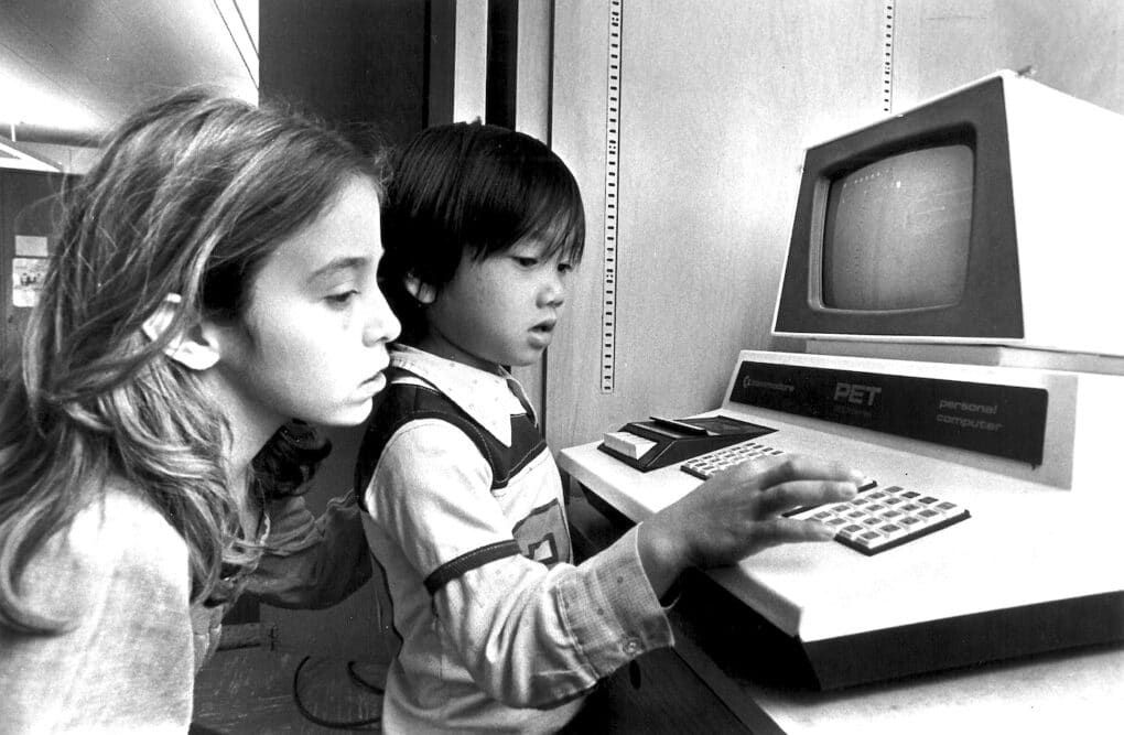 Two children are using an early issue computer. A girl, older, is looking over the shoulder of a younger boy as he types numbers.