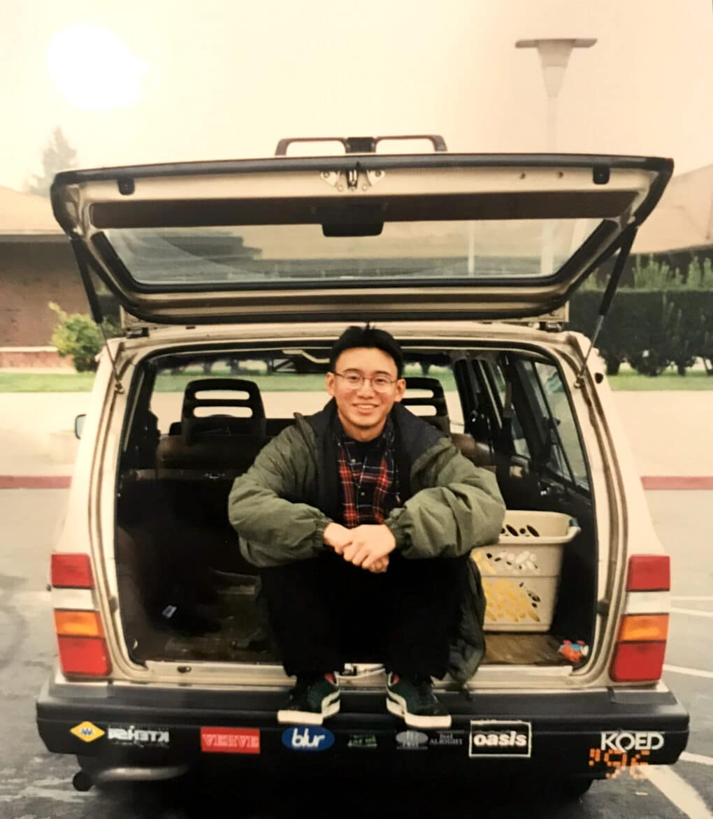 Color vintage photo of smiling young Asian man sitting in the back of a VW sedan with bumper stickers of various music bands including Verve and Oasis.