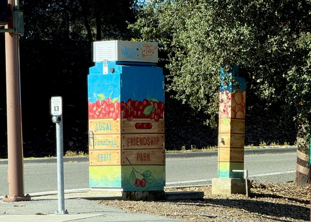 Color photo of a traffic box painted to look like a crate of cherries, and a utility meter painted to look like a (very thin) crate of peaches.