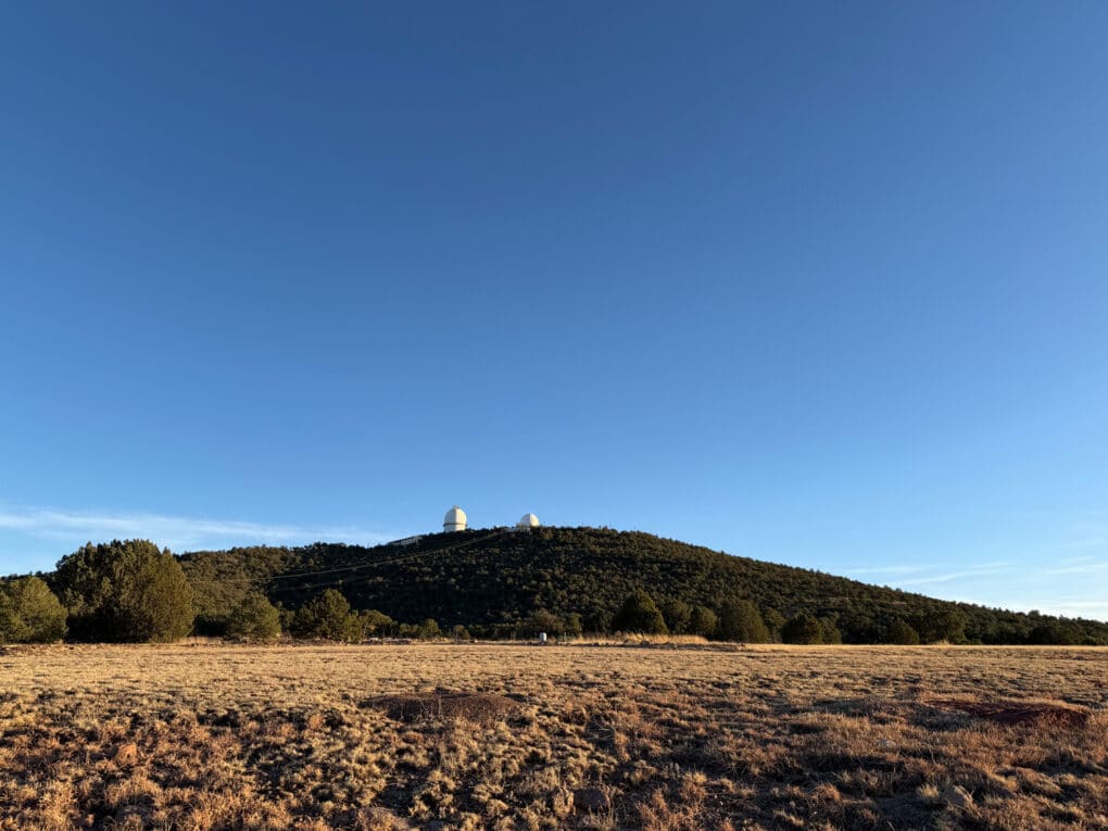 Color photograph showing two white domes of an observatory atop a green hill against a bright blue sky.