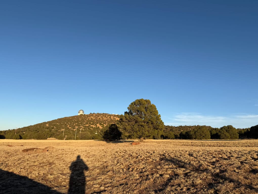 Color photograph of green hill under blue sky. On the crest of the hill is the dome of an observatory. In the foreground is the shadow of the photographer.