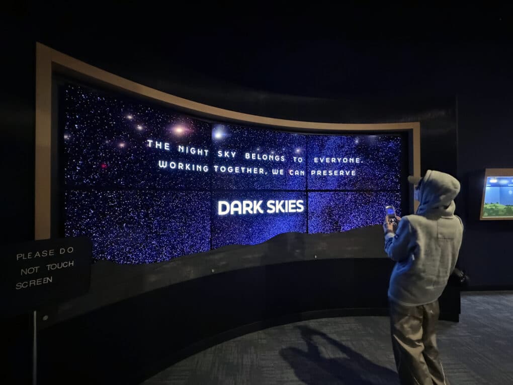 Color photograph of wall-sized lightbox in a museum exhibit, reading "The night sky belongs to everyone. Working together, we can preserve DARK SKIES." A visitor wearing a hoodie and baseball cap is photographing the installation with his phone.