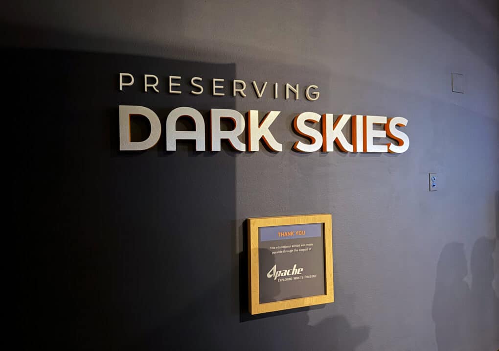 Color photograph with the words PRESERVING DARK SKIES on a gray-painted wall, with a gold-framed sign in which the words "Thank You" in red and "Apache" in white are legible.