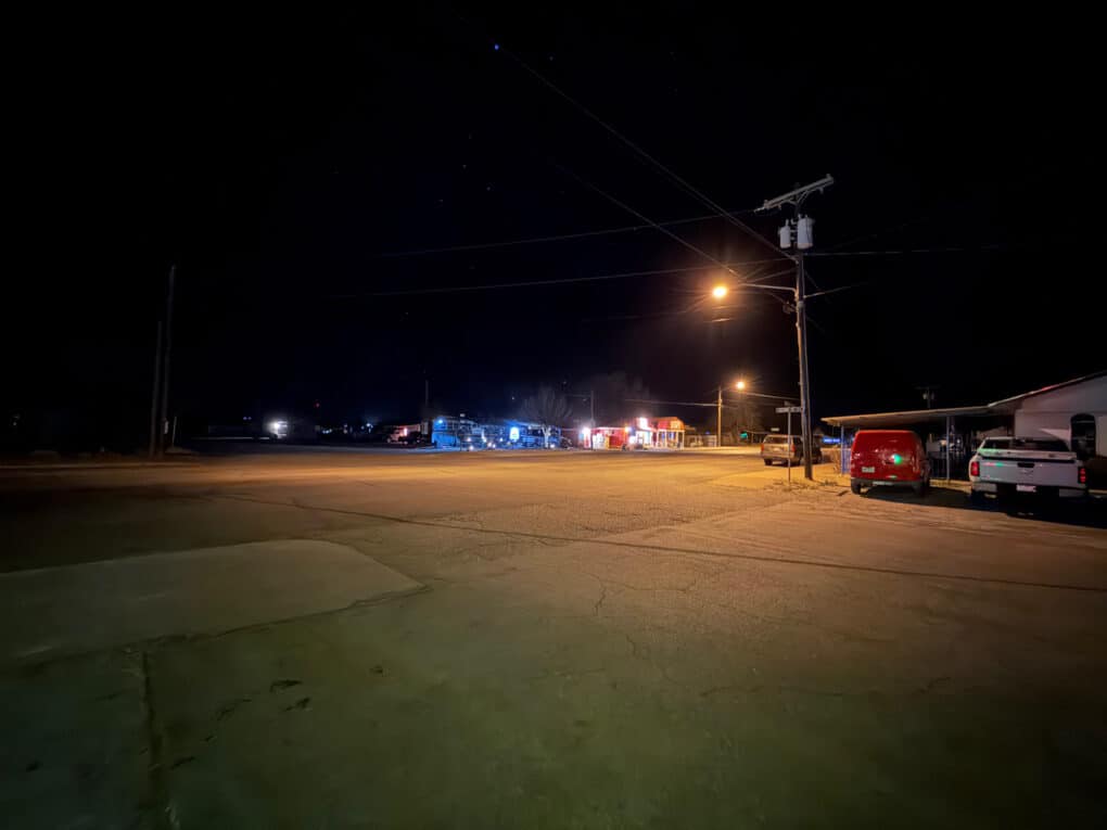 Color photograph of small-town gas station at night, surrounded by dark sky and unlit paved lot.
